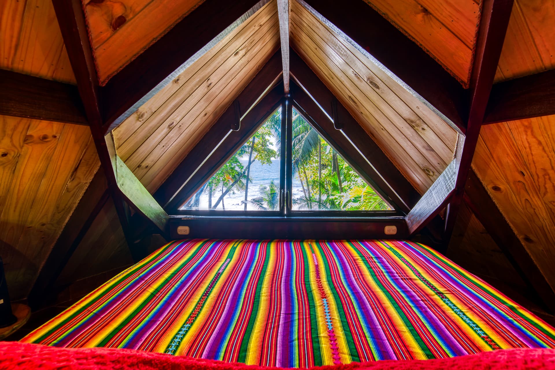 Bed with brightly striped bedding in loft with hardwood vaulted ceilings and a triangle shaped window at foot of bed