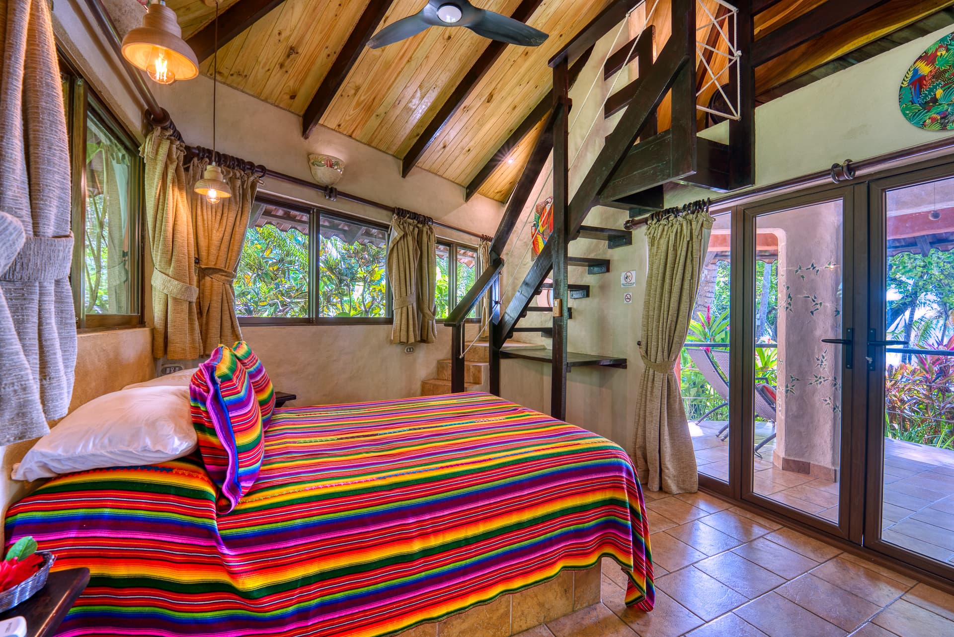 Main floor of bungalow with brightly adorned bed, steps going up to loft, and sliding doors to private patio