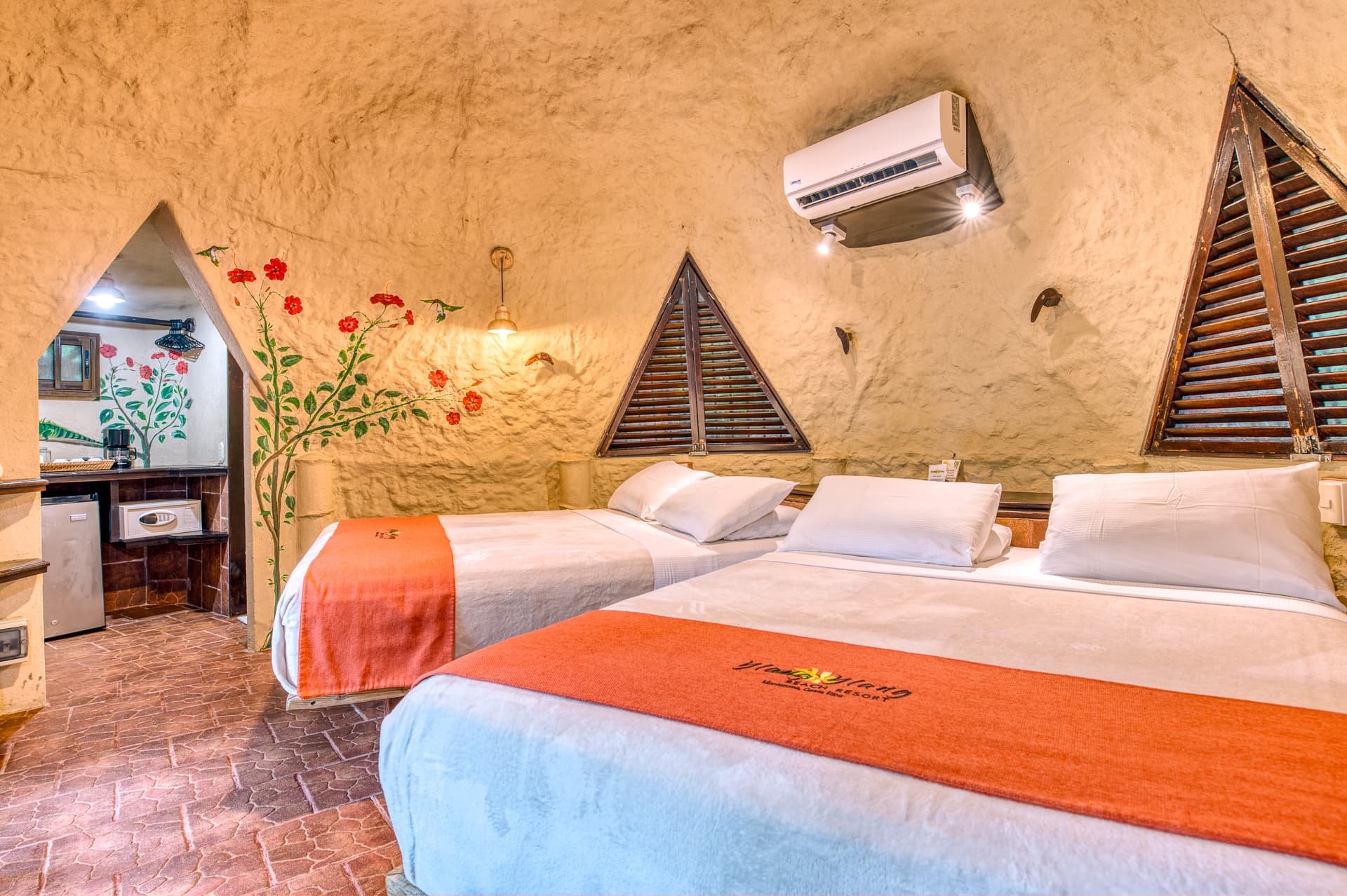 Colibri Bungalow bedroom with two queen beds made with white sheets and orange blankets and doorway leading to bathroom