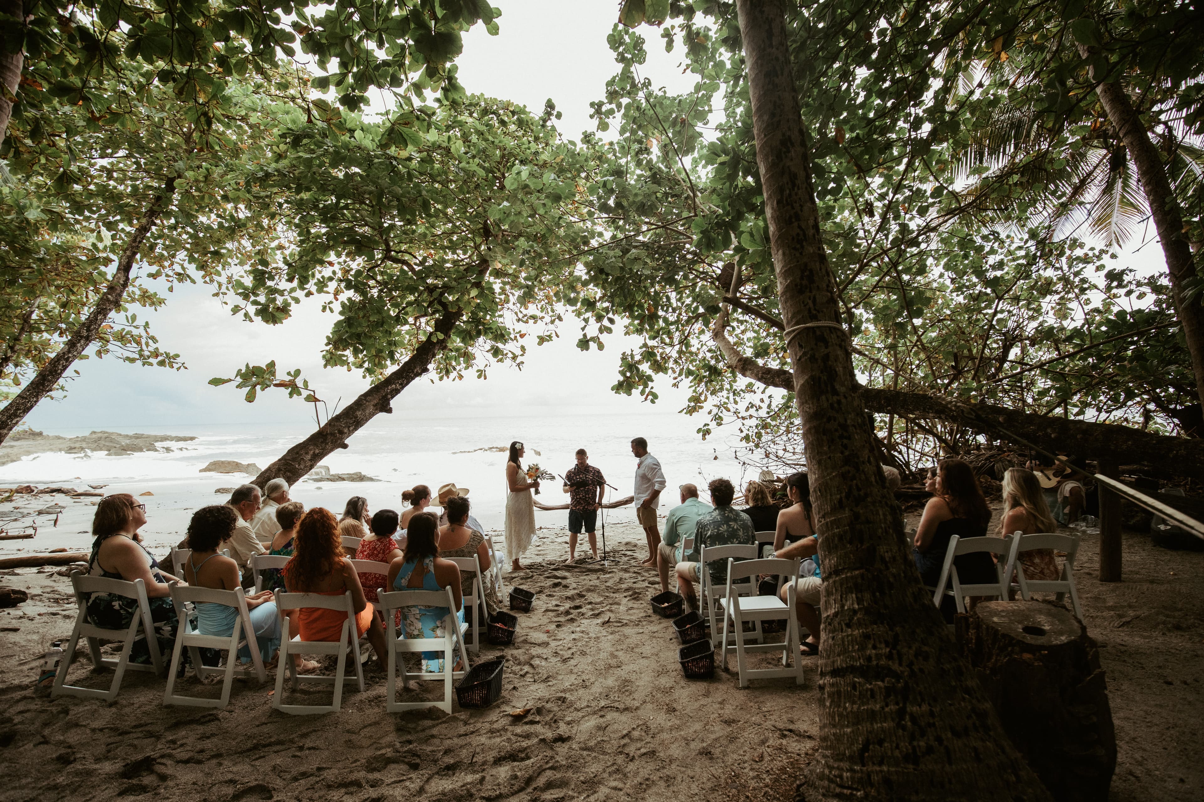 Wedding guests seated in chairs on a tropical beach as an officiant speaks to a bride and groom