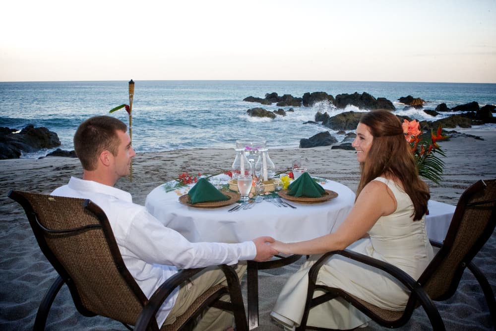 man and woman holding hands at white linnen dinner table on beach by ocean .