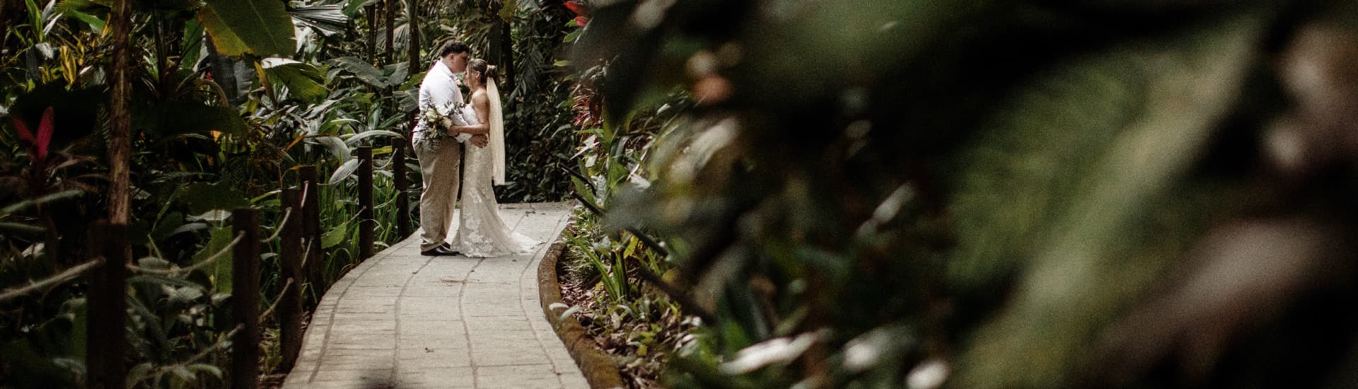 A bride and groom dressed in wedding attire on a stone path surrounded by lush gardens and flowers
