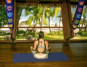 A person meditating on a yoga mat indoors with a tropical beach view.