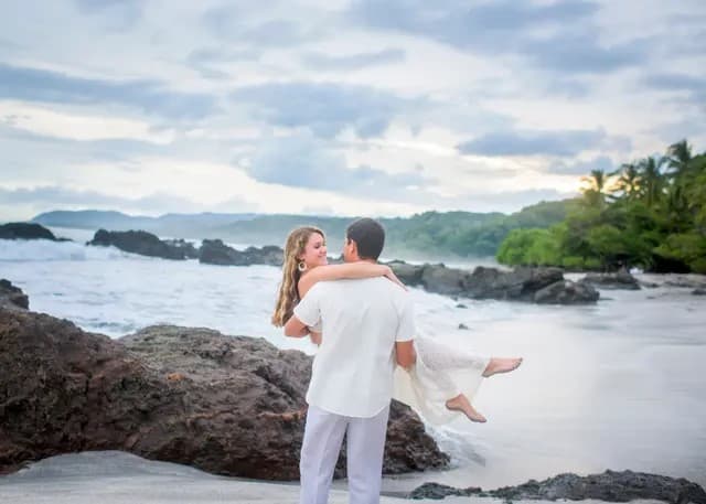 A couple embraces on a rocky beach under a cloudy sky.