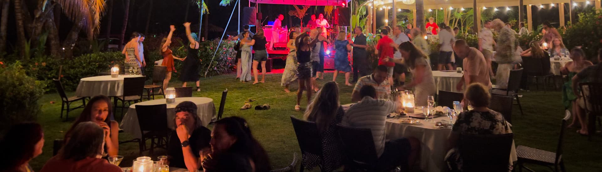 Nighttime scene of a live band performing while some people dance and others eat and talk at candlelit tables