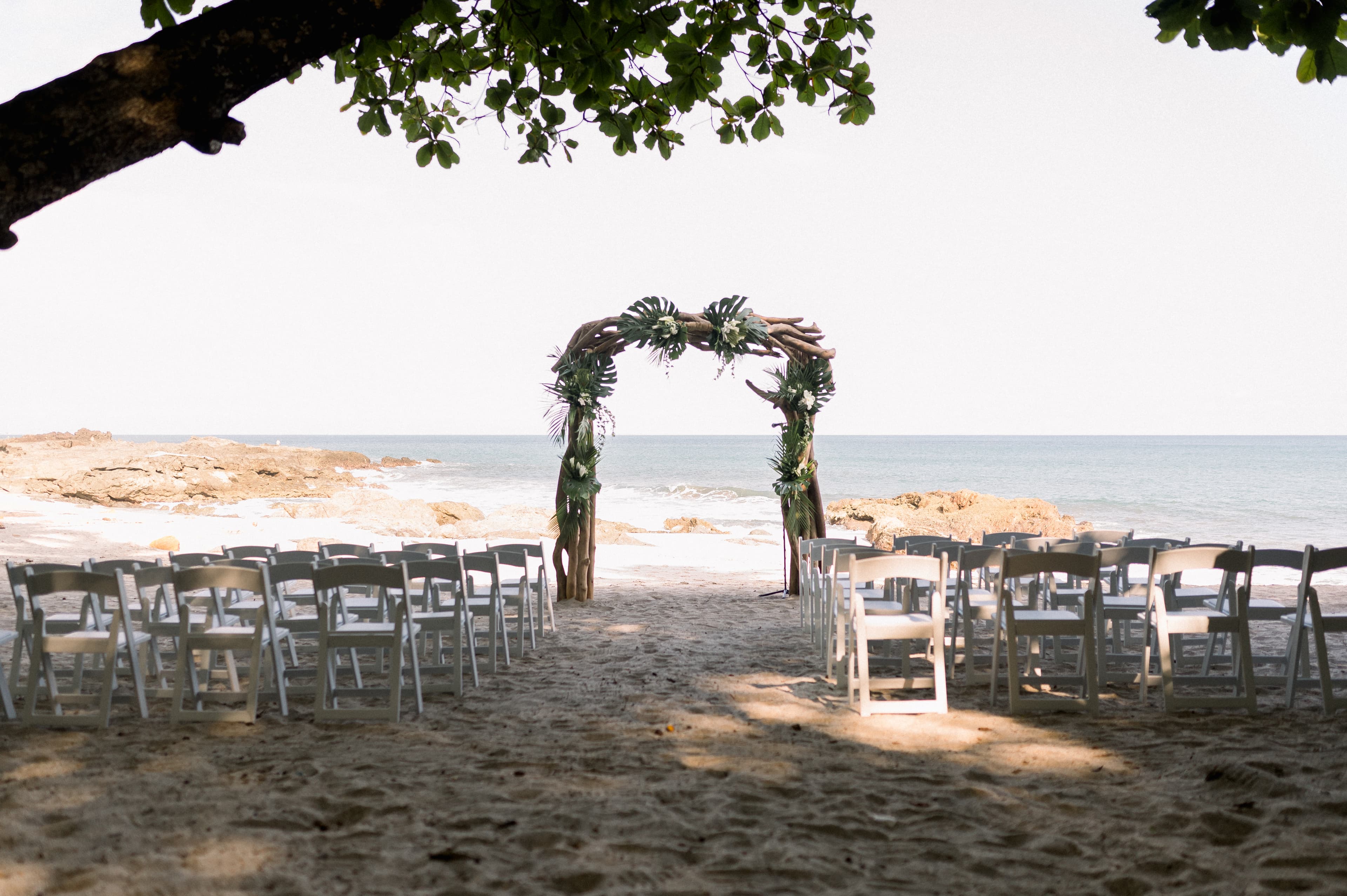A sandy beach near the ocean is set up for a wedding ceremony under palm trees with a driftwood arch decorated with tropical foliage and rows of chairs for guests