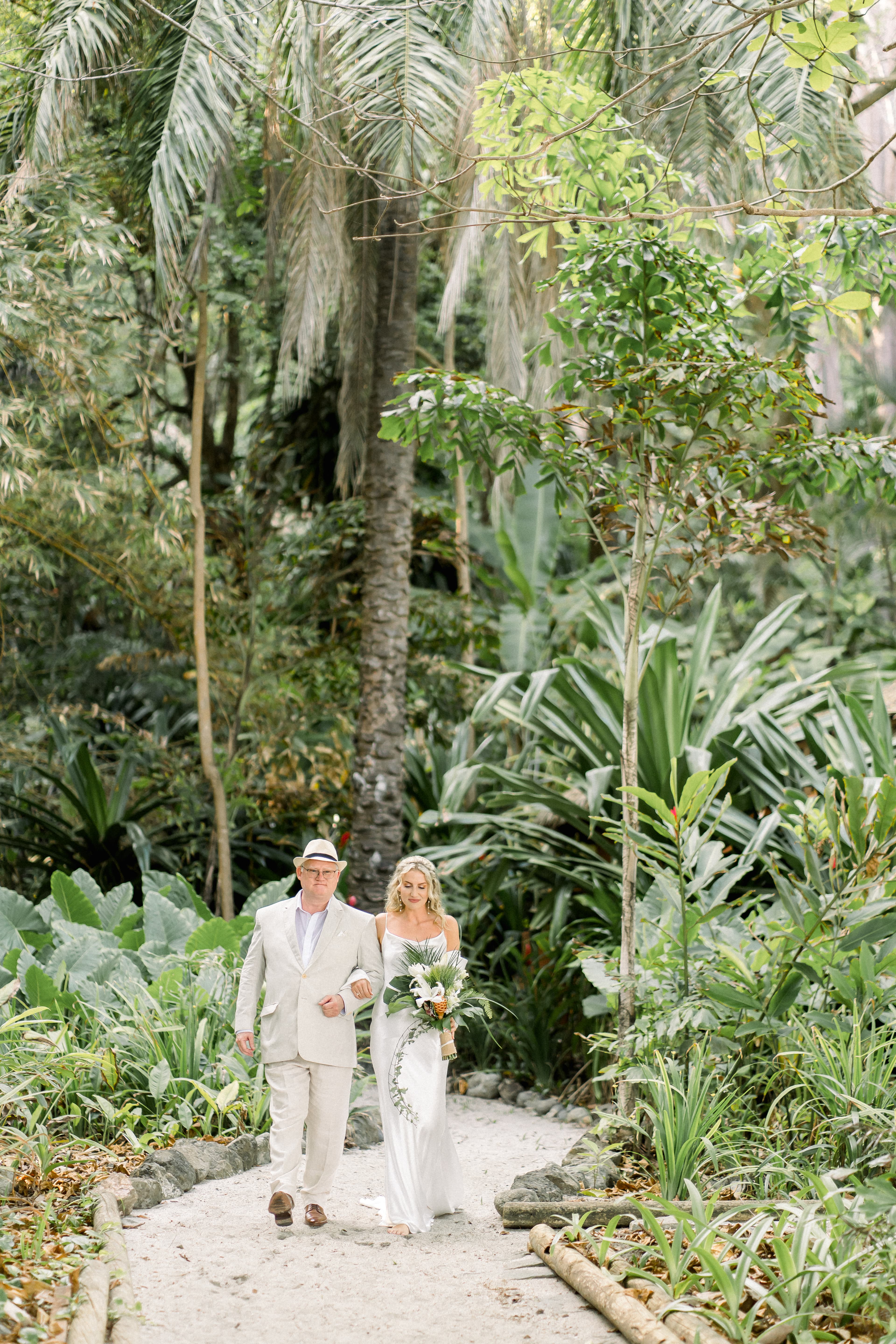 A bride and groom strolling arm in arm down a meandering pathway surrounded by lush tropical foliage. The bride is carrying an impressive bouquet of tropical flowers.