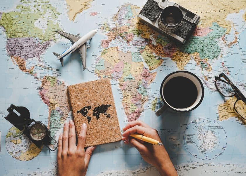 A hand holds a notebook on a world map surrounded by a coffee cup, camera, airplane model, and compass. A hand holds a notebook on a world map surrounded by a coffee cup, camera, airplane model, and compass.