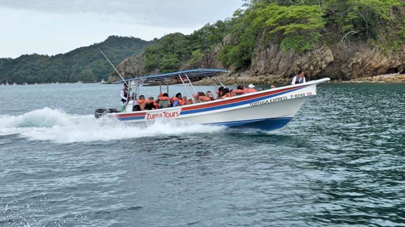 A speedboat named "Zuma Tours" carries passengers through the ocean near a rocky coastline.