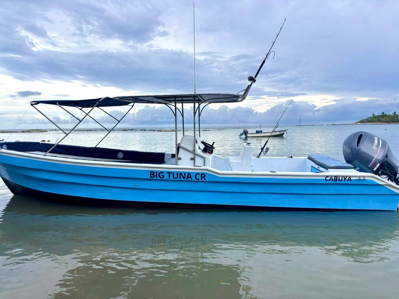 A blue fishing boat named "BIG TUNA CR" is anchored in calm waters under a cloudy sky.