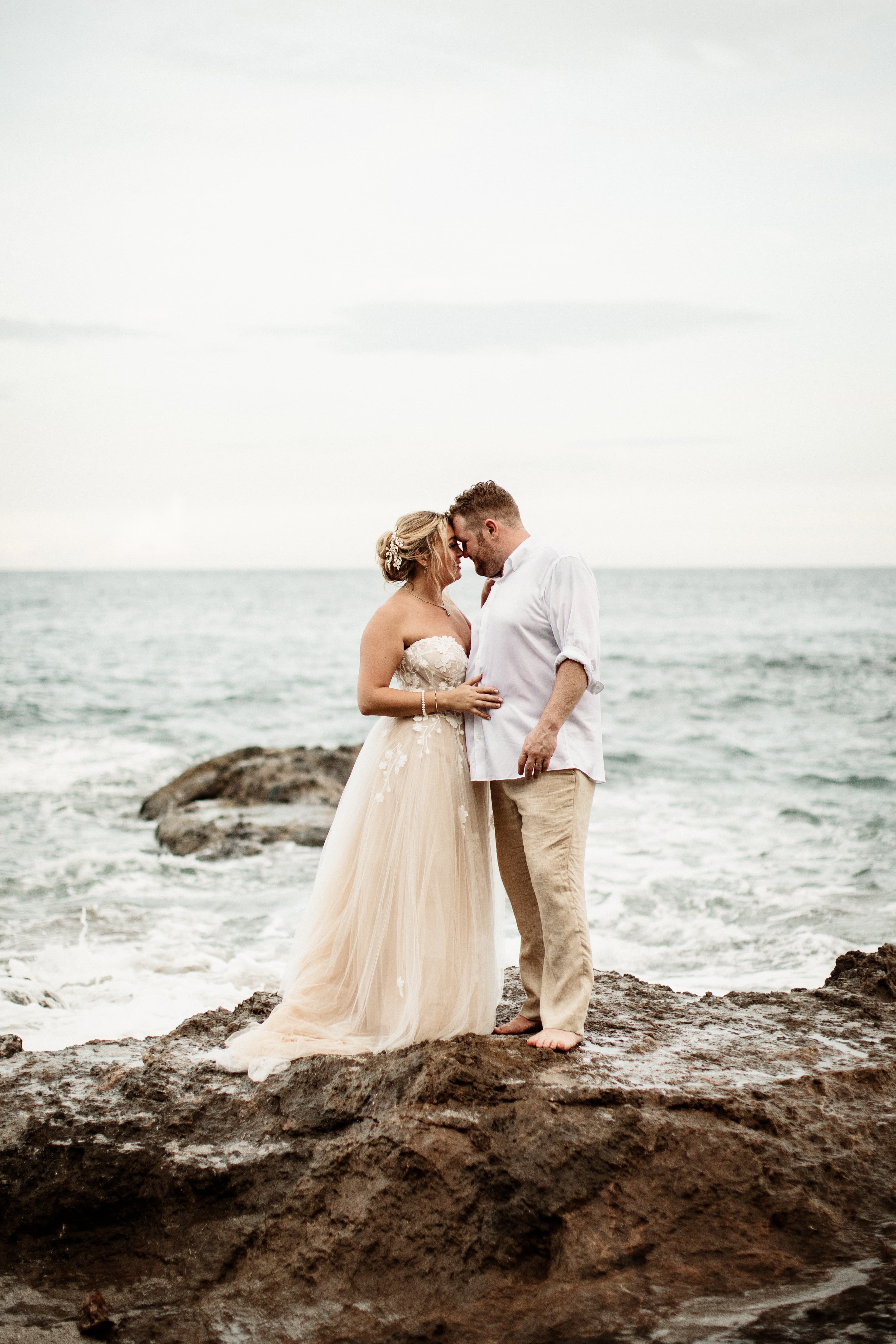 A bride and groom stand on a large rock by the ocean's shore, while embracing with their foreheads touching