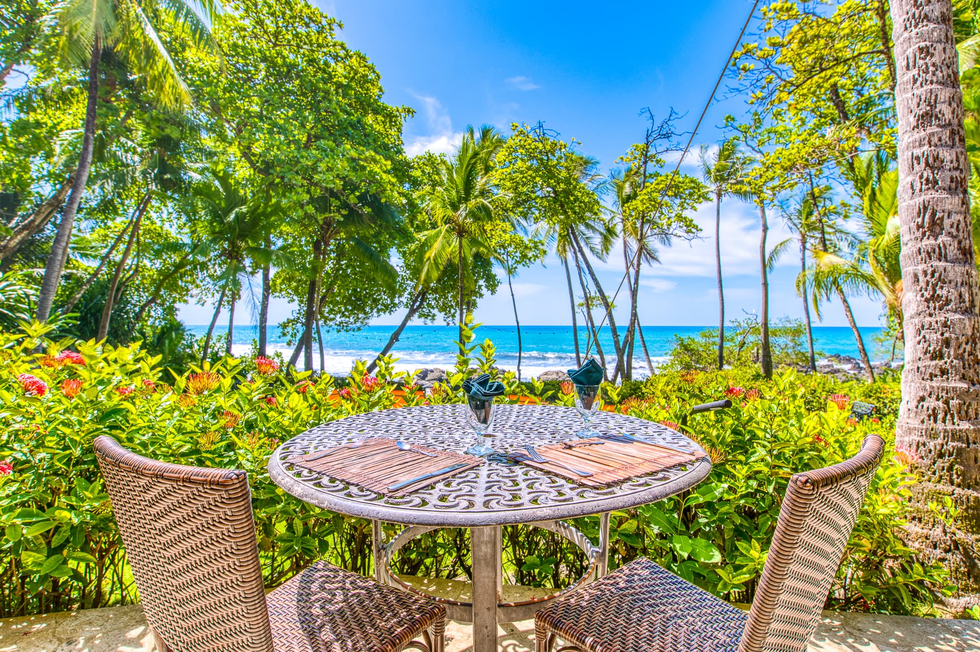 A seaside table set for two, surrounded by lush greenery and view of the ocean.