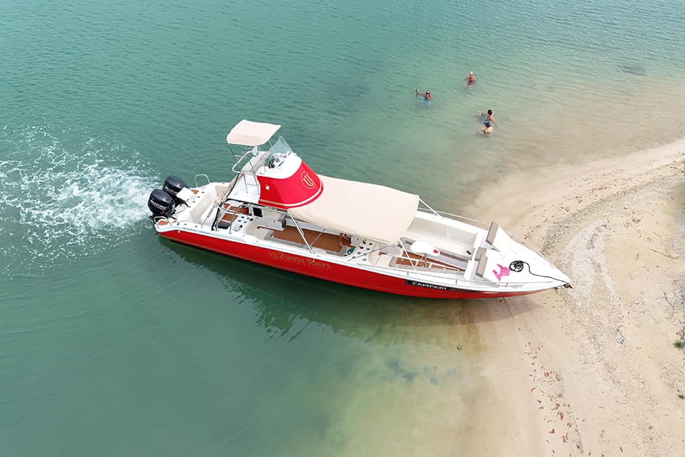 A red and white boat named "Zuma Tours" is moored near a sandy shore with people swimming nearby.