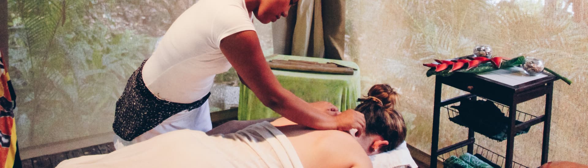 A female masseuse in a tropical cabana massaging the neck of a woman who is draped in a sheet and lying face down on a massage table