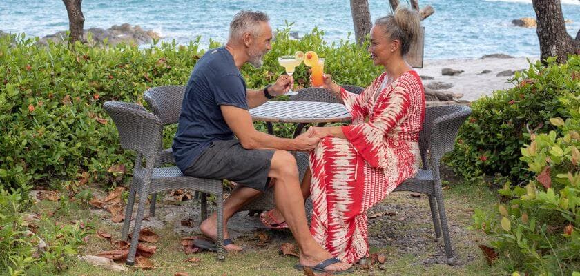 A couple enjoys tropical drinks at a table by the beach amidst lush greenery.