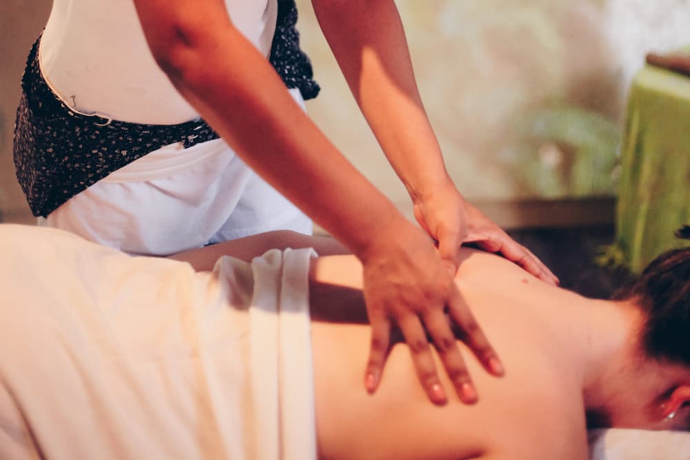 A masseuse's hands massaging the back of a woman lying face down on a massage table, draped in a white sheet, with her hair in a bun