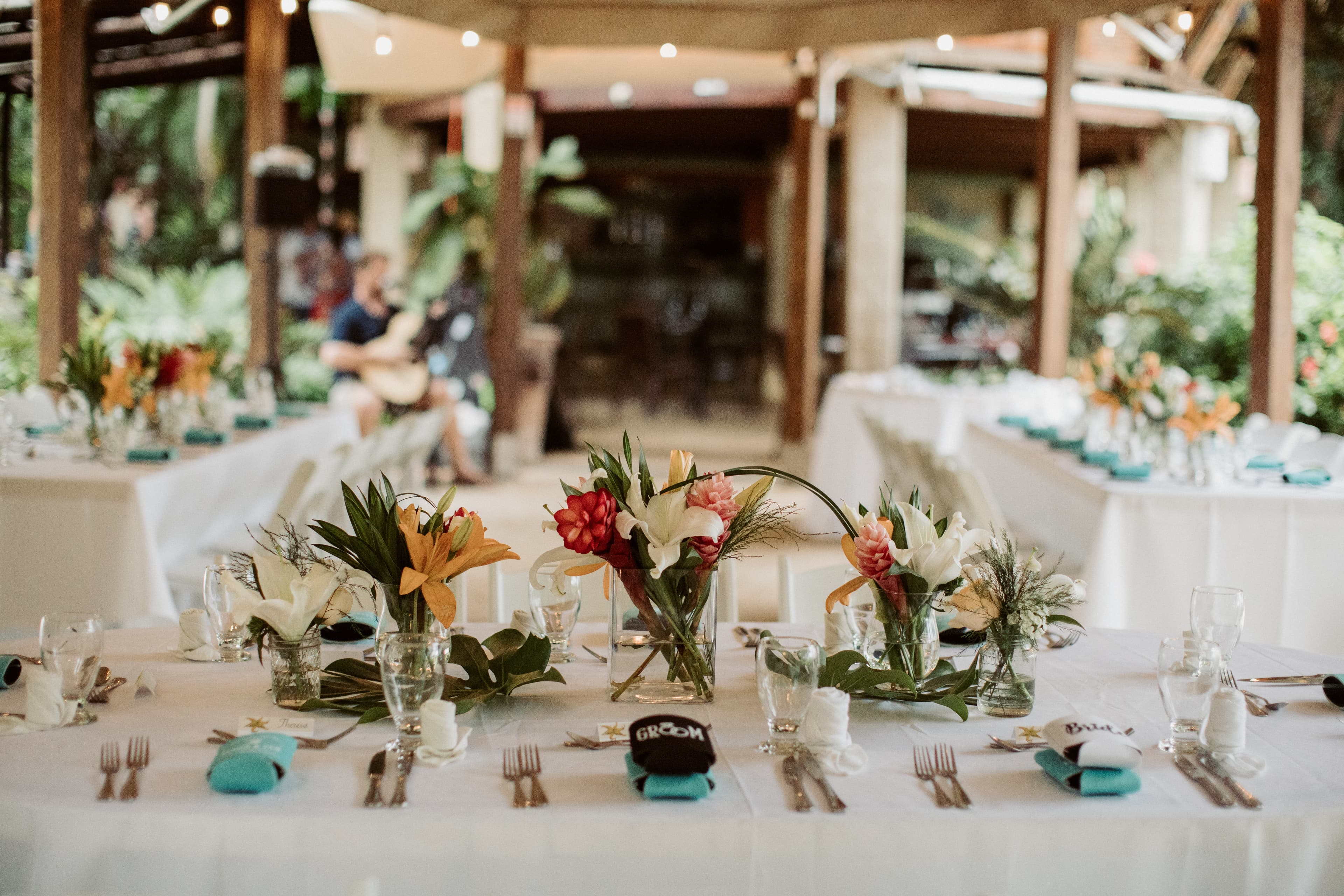 An outdoor wooden pavilion set up for a wedding reception with tables covered in white tablecloths and decorated with tropical floral centerpieces and string lights hanging around the perimeter