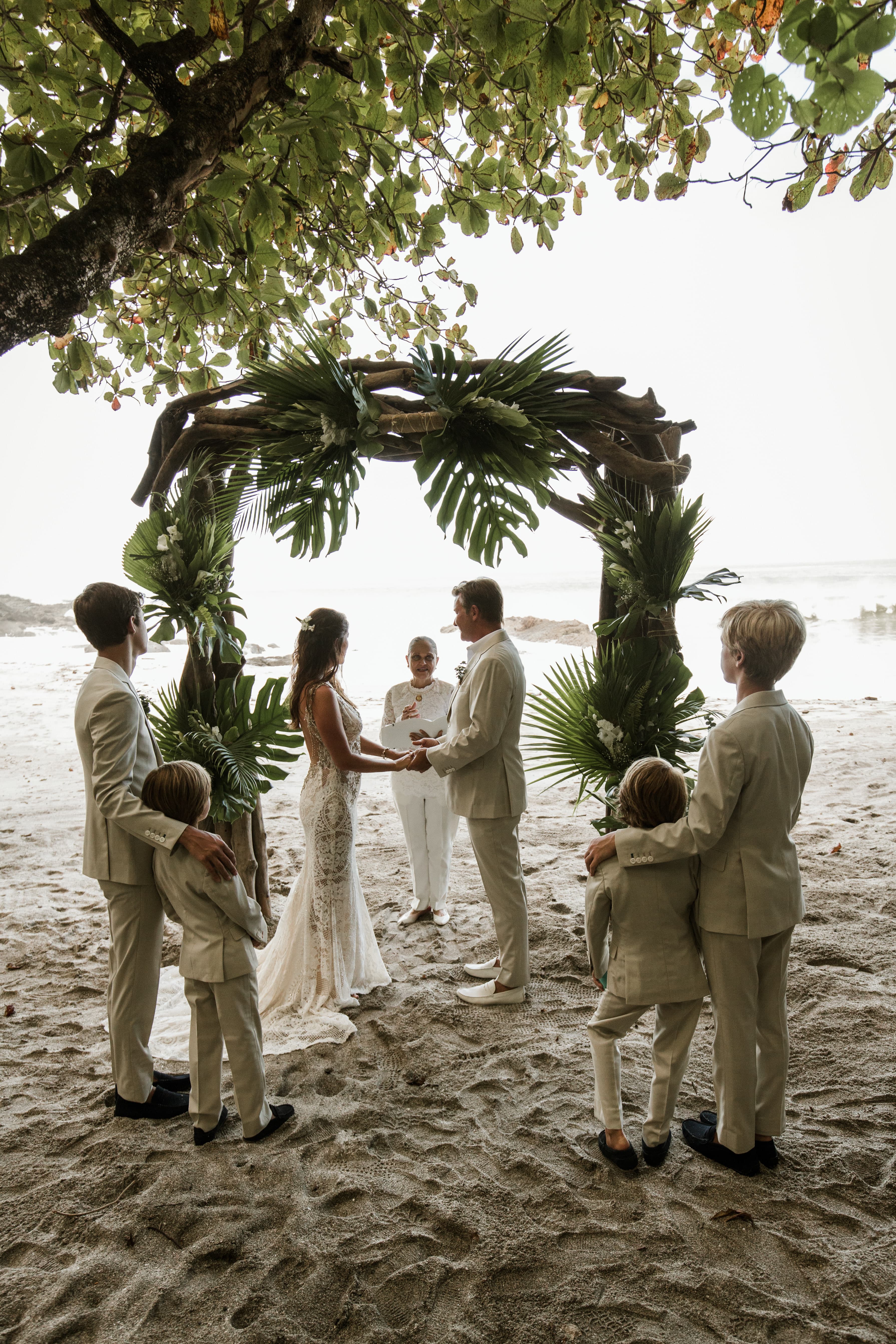 A bride and groom stand on a sandy beach, clasping hands under a driftwood arch decorated with tropical foliage while an officiant speaks and four young boys in matching khaki suits look on
