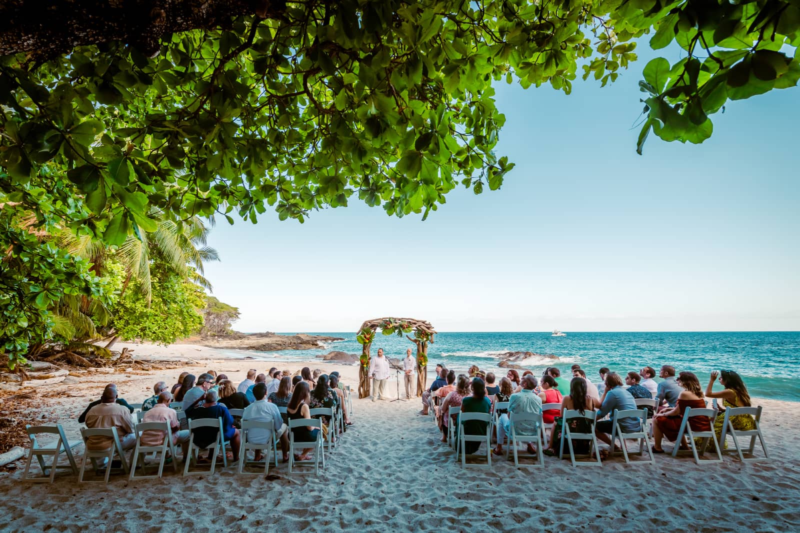 A beach wedding ceremony set under a lush canopy, with guests seated in rows facing the couple and the ocean.
