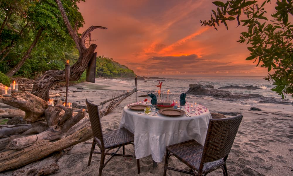 Candlelit table set for two on a sandy beach with a sunset of oranges and lavenders over the ocean