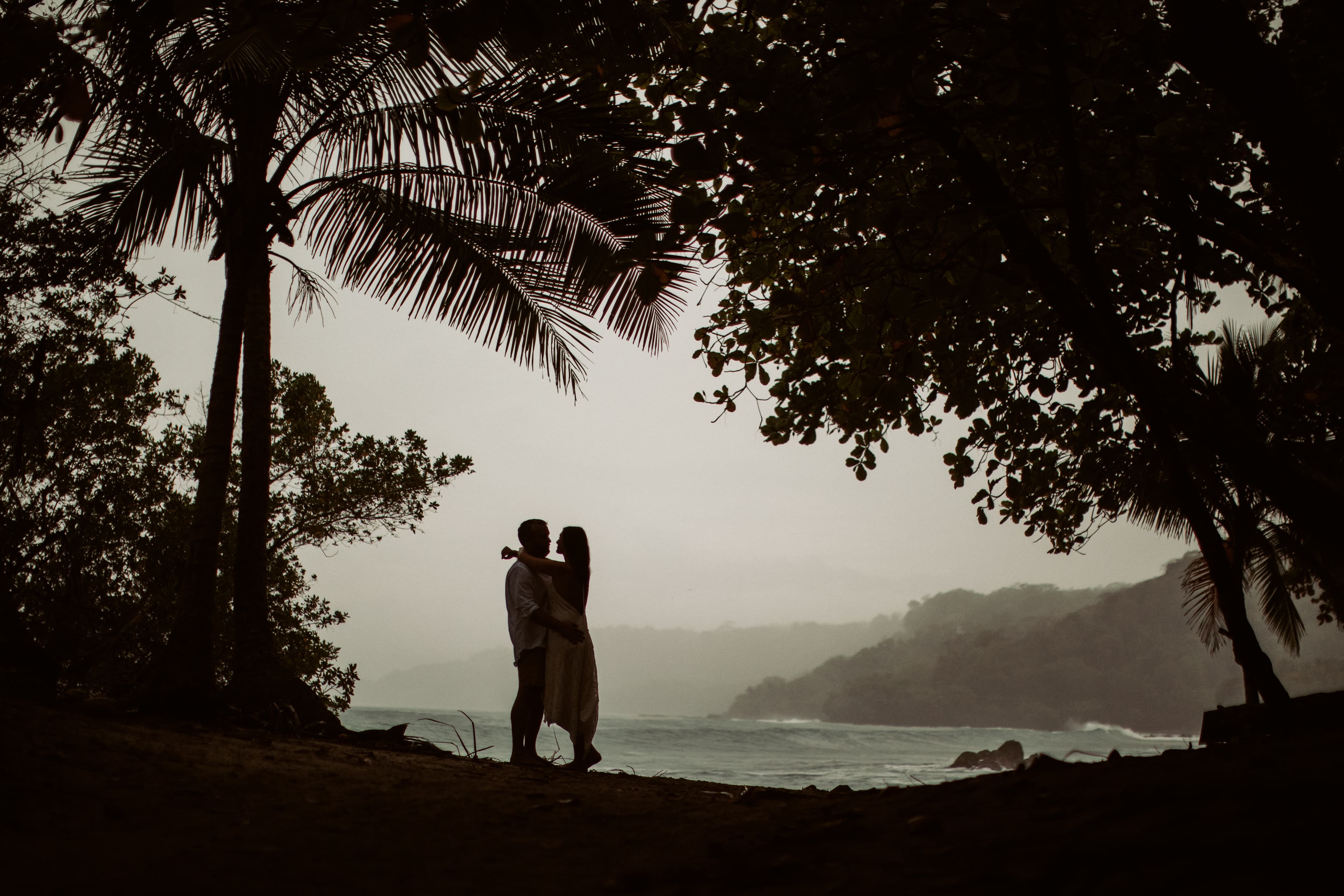 An embracing couple standing under palm trees on a sandy beach and silhouetted against a misty sky and the ocean