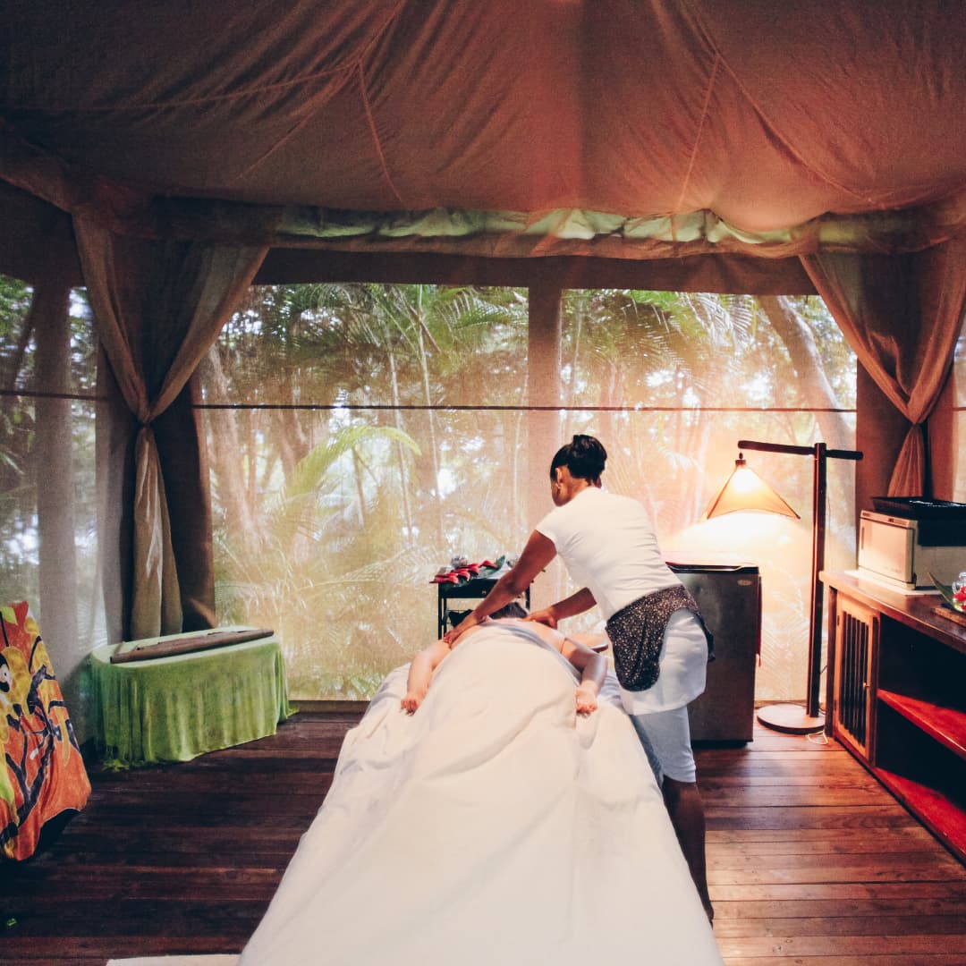 Masseuse in a jungle cabana massaging a person draped in a white sheet lying on a massage table