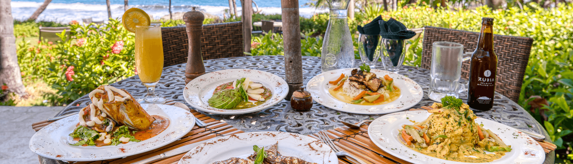 Outdoor wrought iron table with a view of the beach and set with a variety of expertly plated gourmet dishes