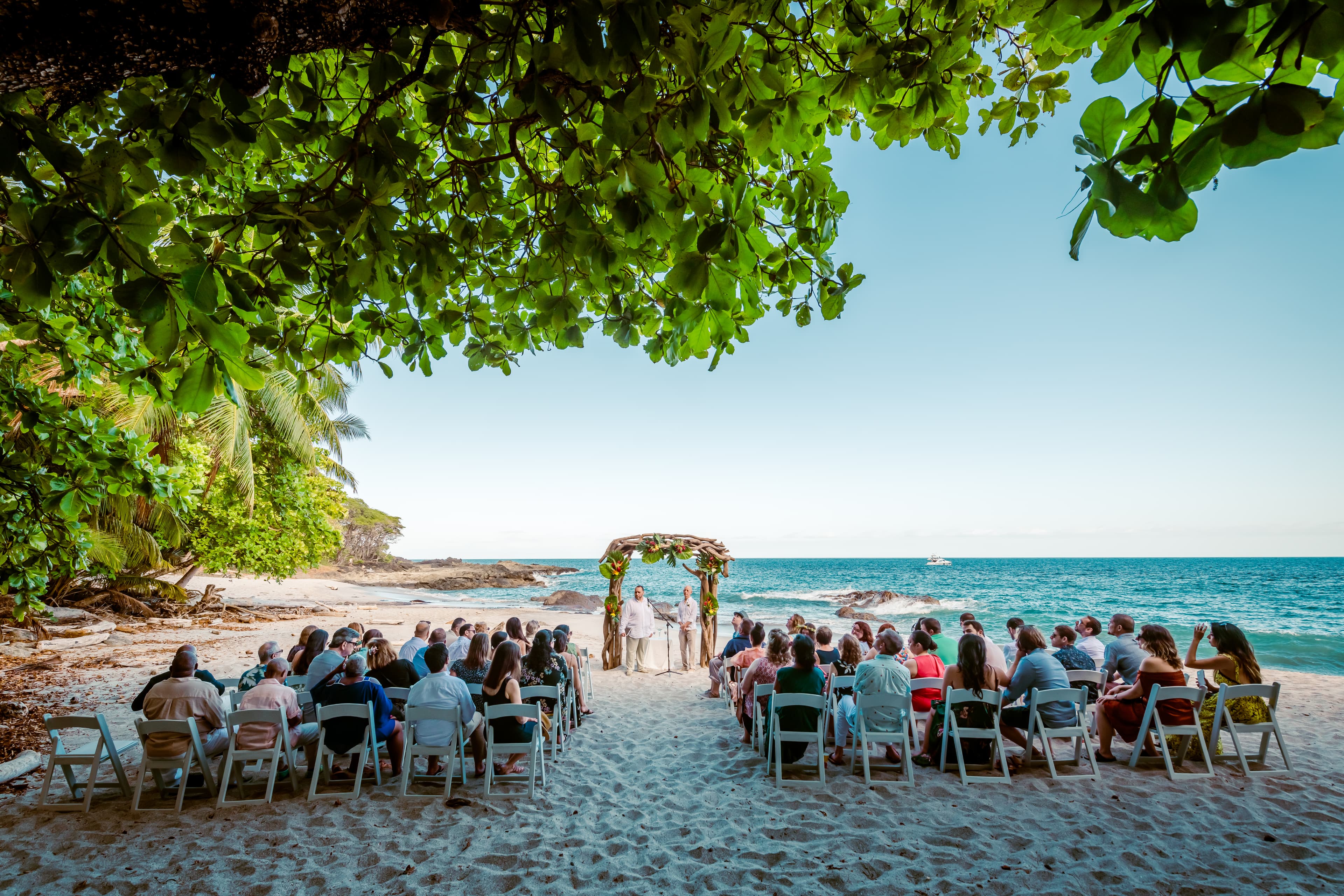 A groom and officiant waiting under a driftwood arch decorated with tropical flowers on a beach with wedding guests seated on chairs looking on