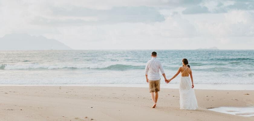 A couple walks hand in hand along a sandy beach, with waves lapping at the shore.