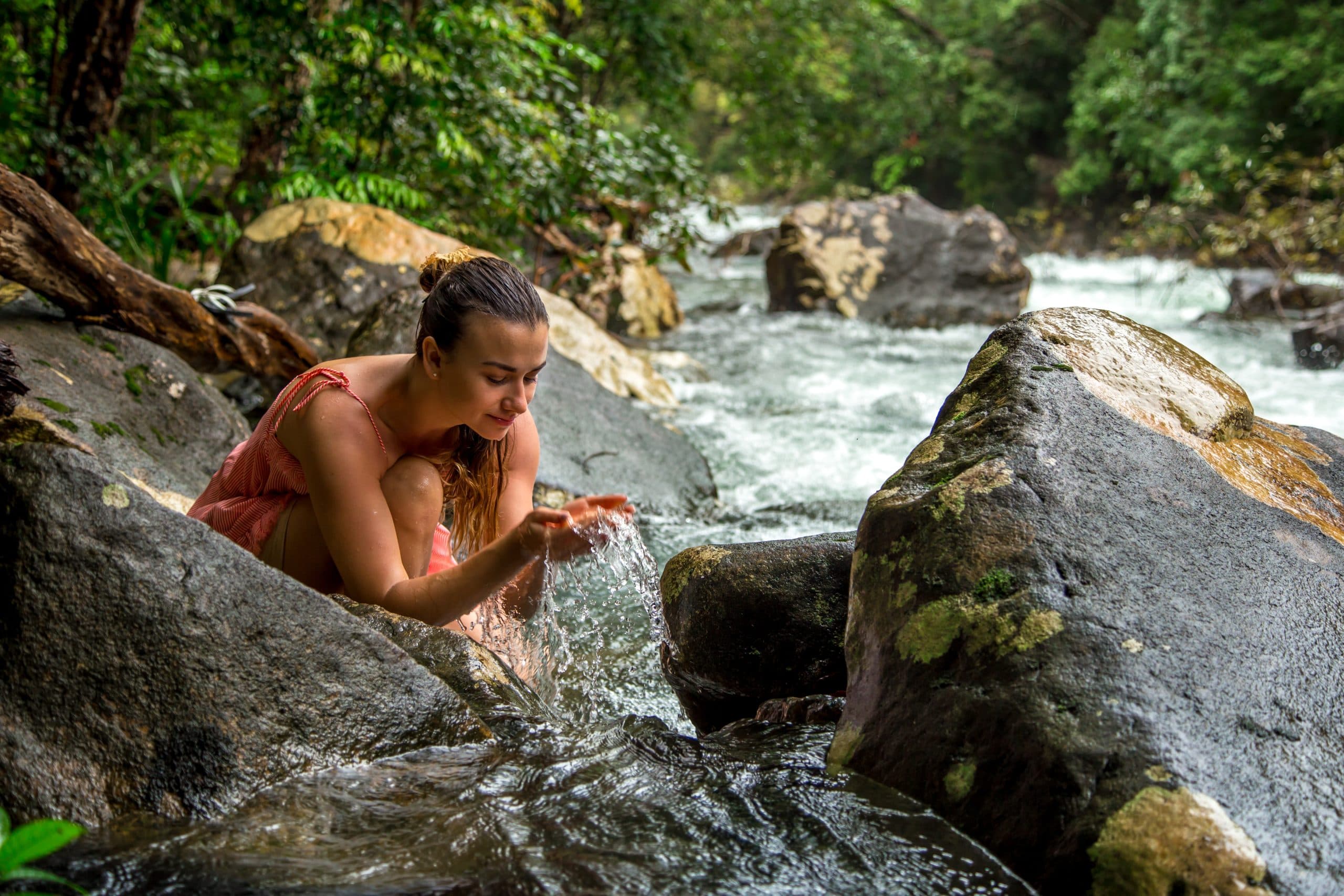 A woman in a pink dress crouches by a river, cupping water in her hands amidst lush greenery. A woman in a pink dress crouches by a river, cupping water in her hands amidst lush greenery.