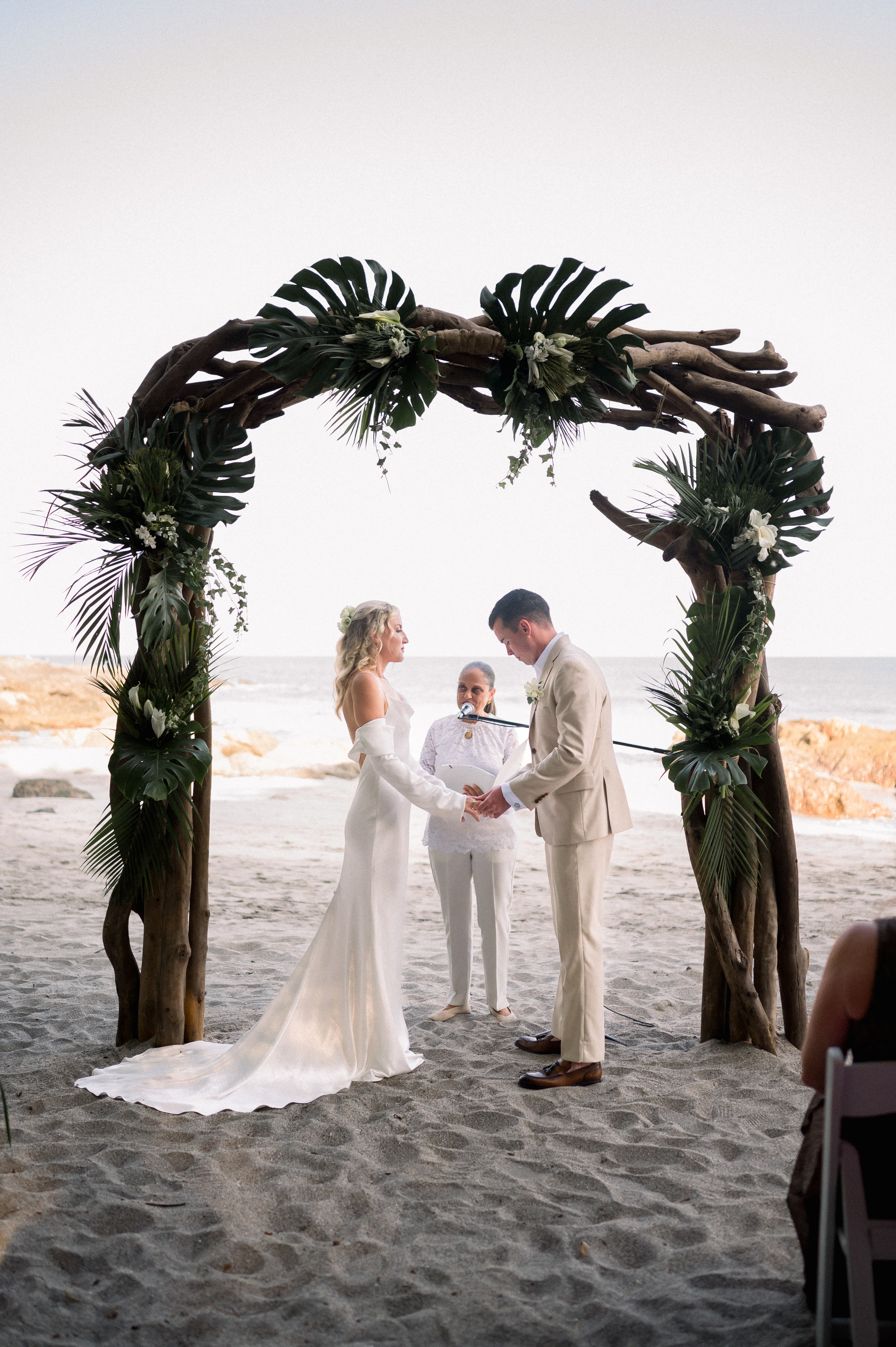 A bride and groom stand on a sandy beach, facing each other with their hands clasped under a driftwood arch decorated with tropical foliage, while an officiant speaks