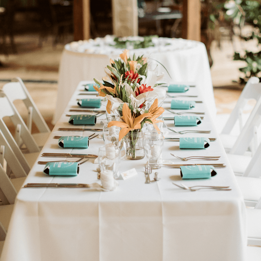 Rectangular table set for wedding party, tropical flowers, candles, place settings on white tablecloth and white wood chairs.