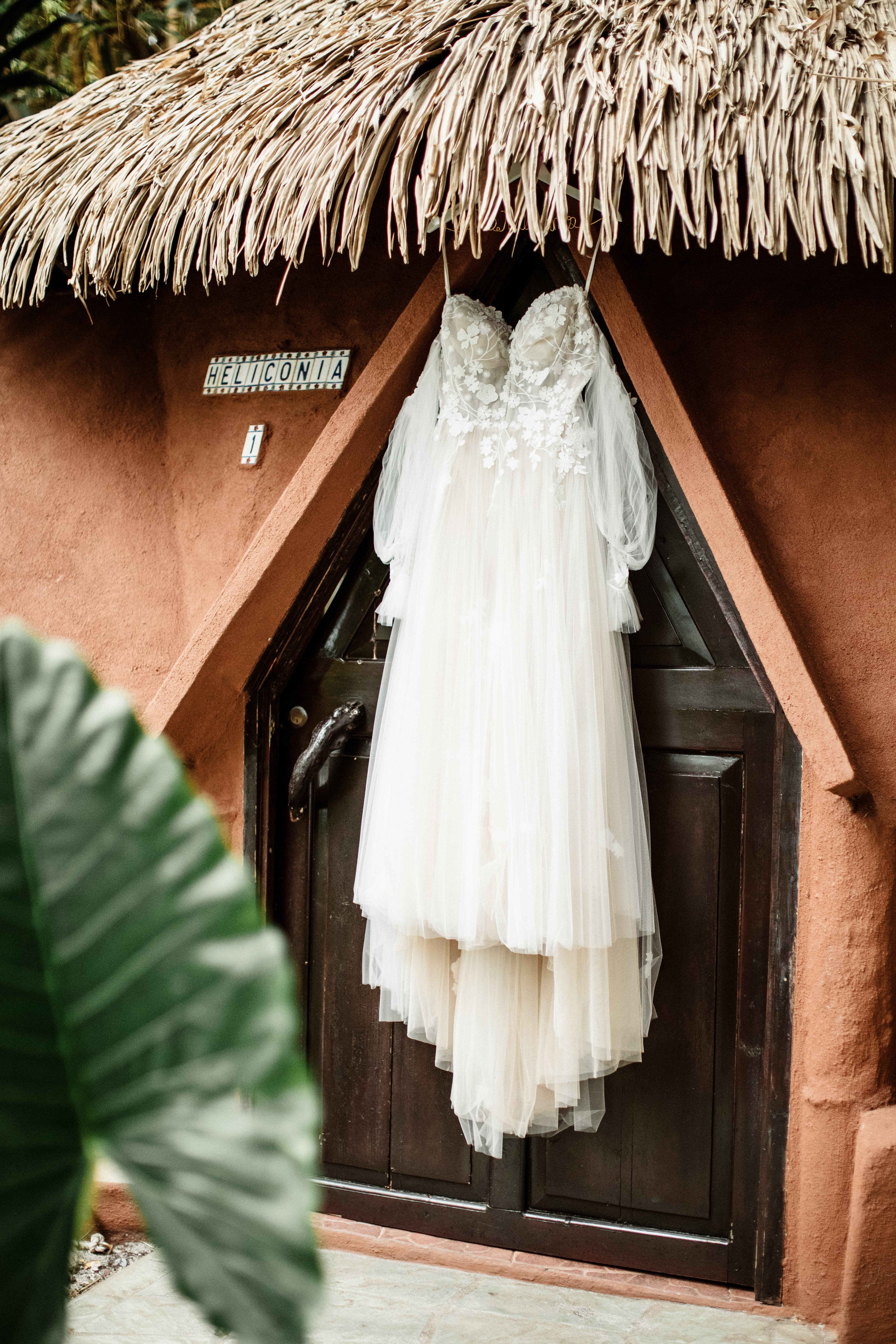 A white wedding dress hanging on the exterior door of a cabana with a thatched roof