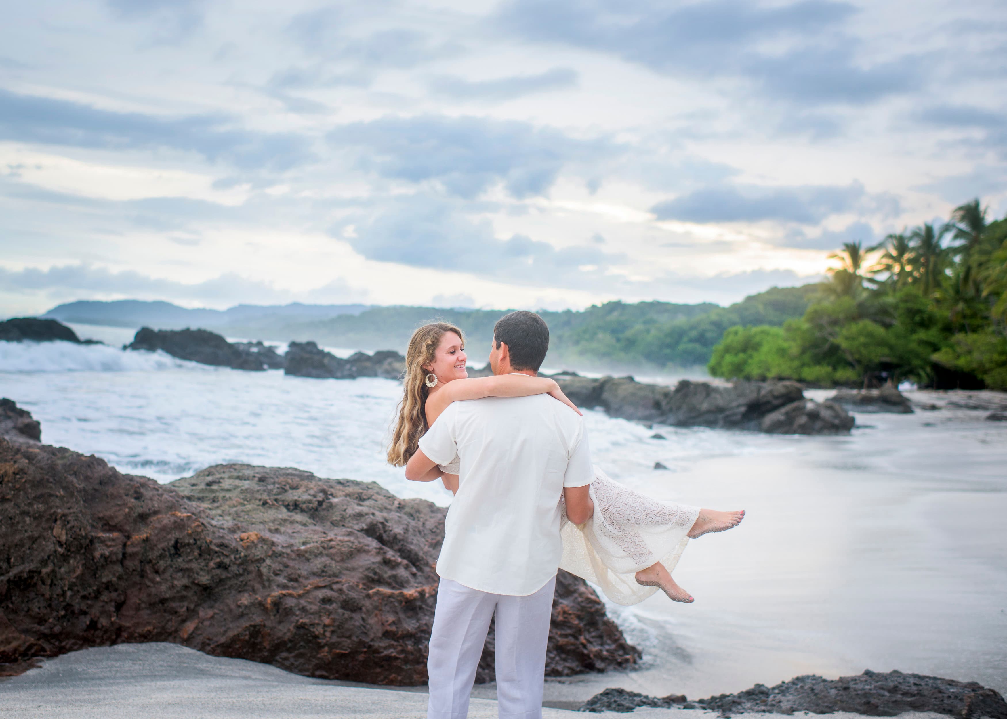 A woman barefoot and dressed in white being held in the arms of a man dressed in white on a rocky coast with waves crashing around them
