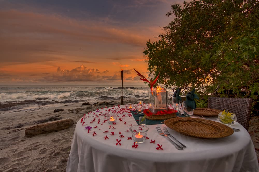 white linen romantic table with flowers, candlelight on beach with ocean in background.