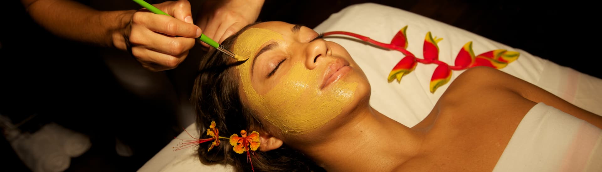 A woman draped in a sheet lying face up looking blissful with her eyes closed while her face is being brushed with a creamy facial treatment