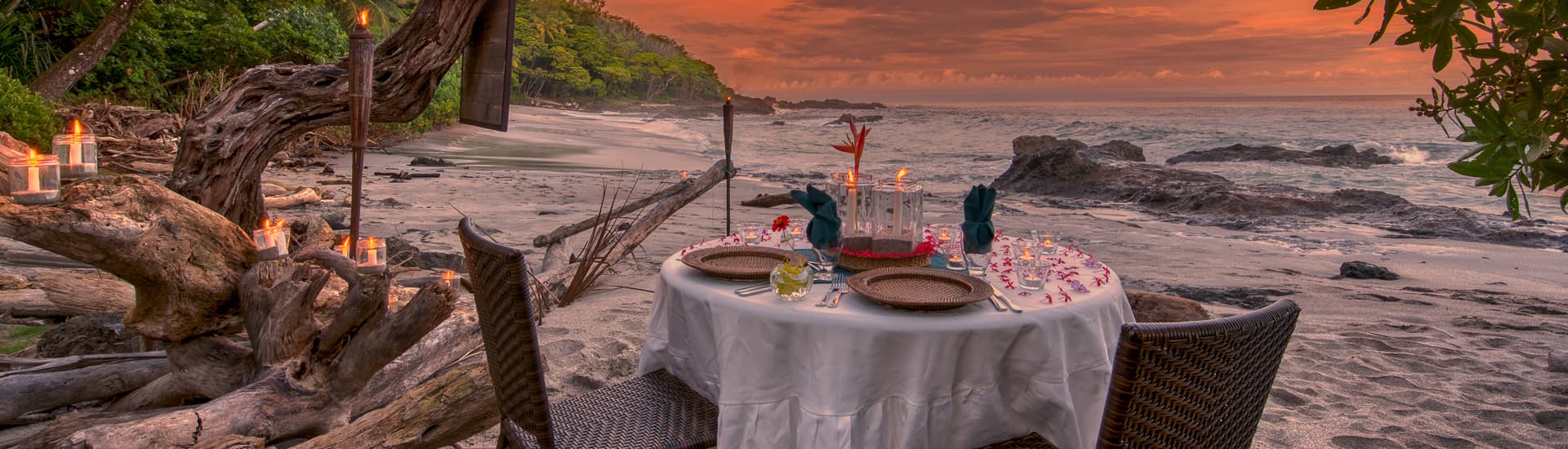 Dining table on beach with table cloth, driftwood and ocean in the background with the sun setting and the jungle off shore