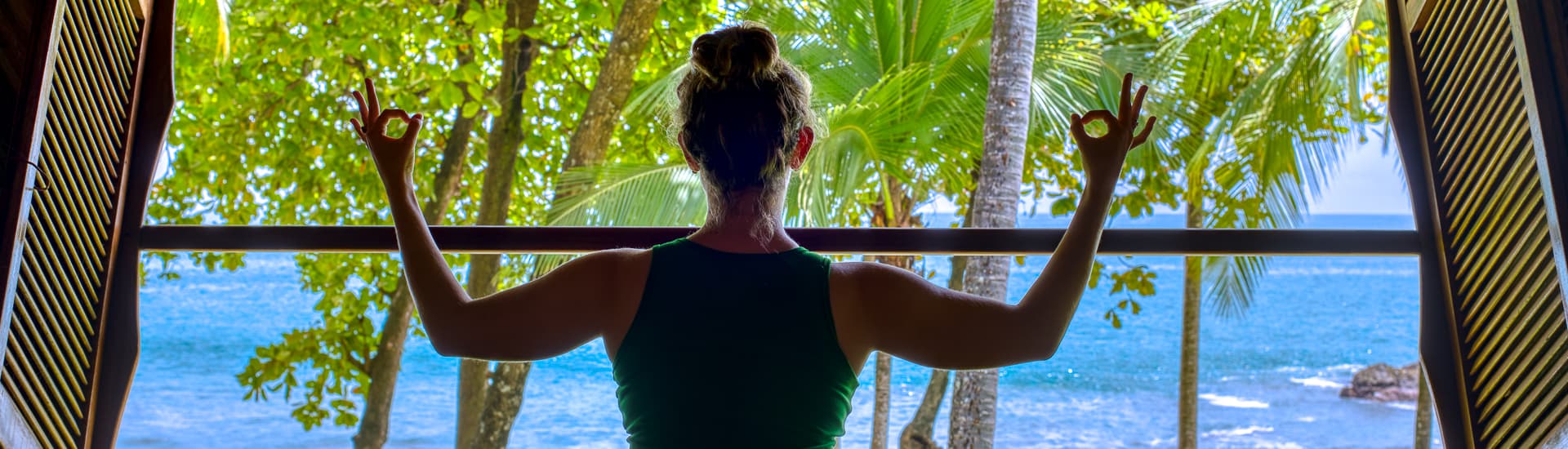 woman in yoga post inside building looking out at ocean with palm trees nearby