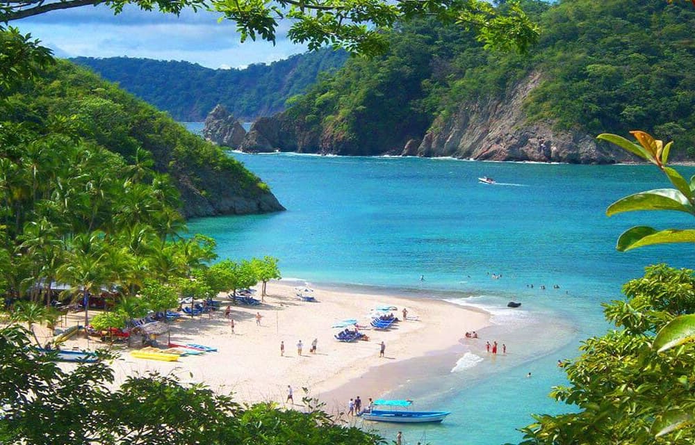 aerial photo above sandy beach with people in water, boats and kayaks and jungle and mountains all around.