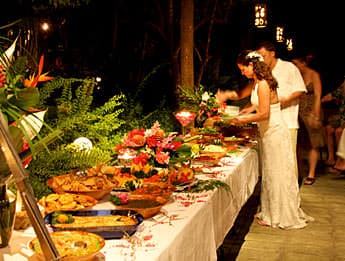 A bride serves food from a beautifully arranged buffet table adorned with tropical flowers and greenery.
