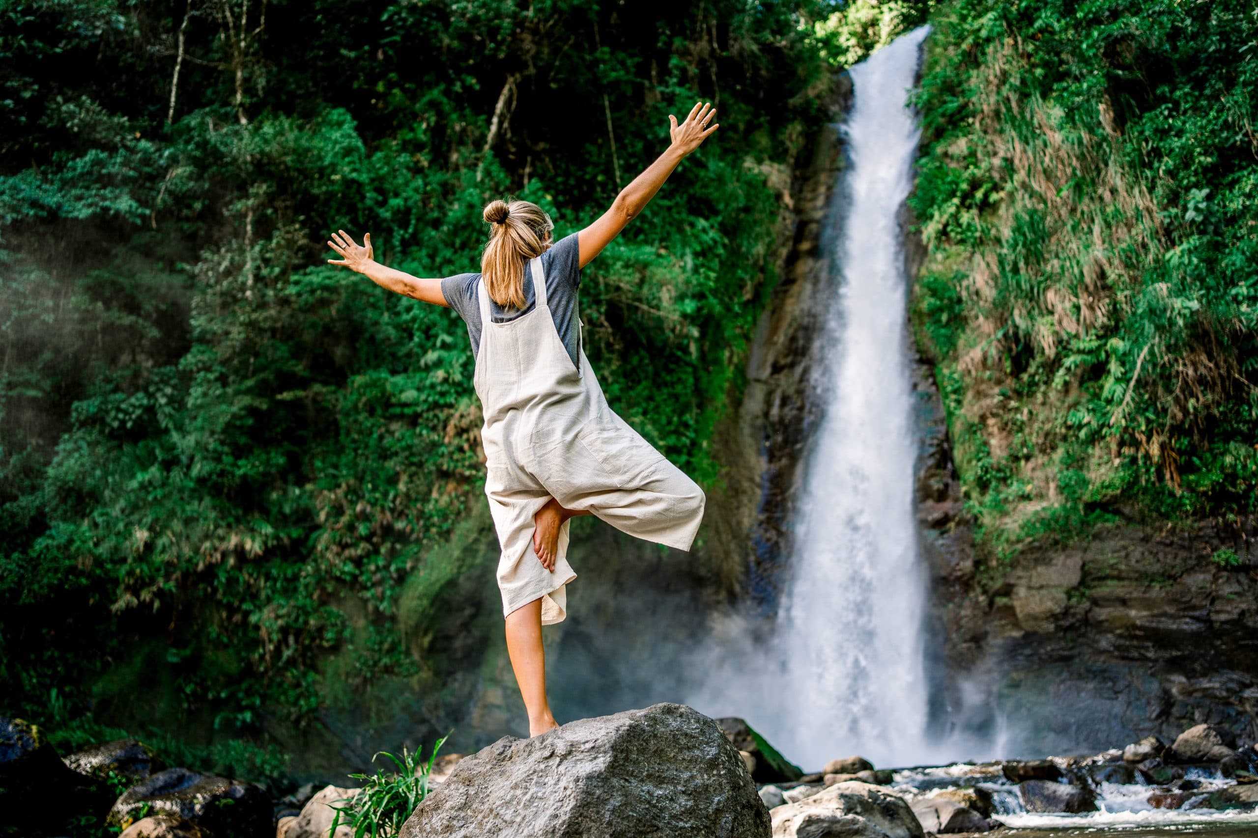 A person balances on a rock with arms outstretched near a waterfall surrounded by lush greenery. A person balances on a rock with arms outstretched near a waterfall surrounded by lush greenery.