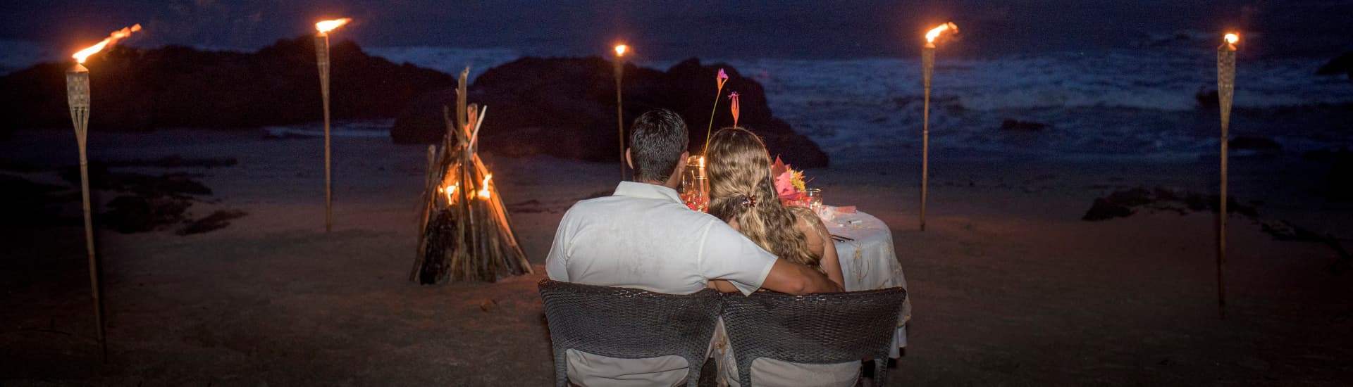 Couple sitting with their backs to the camera at a candlelit table surrounded by lit tiki torches on the beach looking out to the ocean