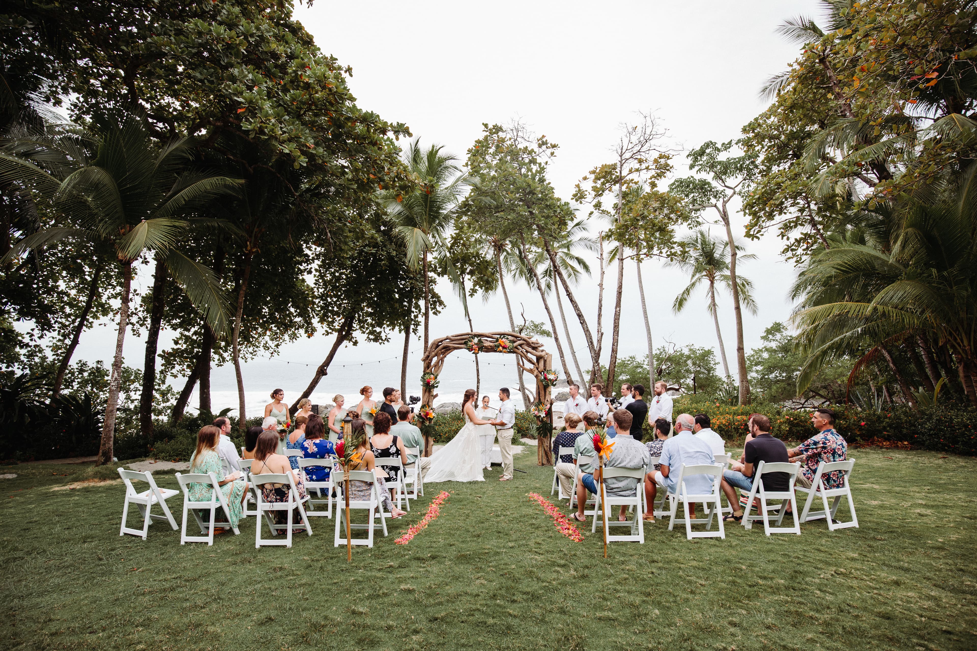 Wedding guests seated in chairs on a tropical beach watching a bride and groom with their hands clasped under a driftwood arch
