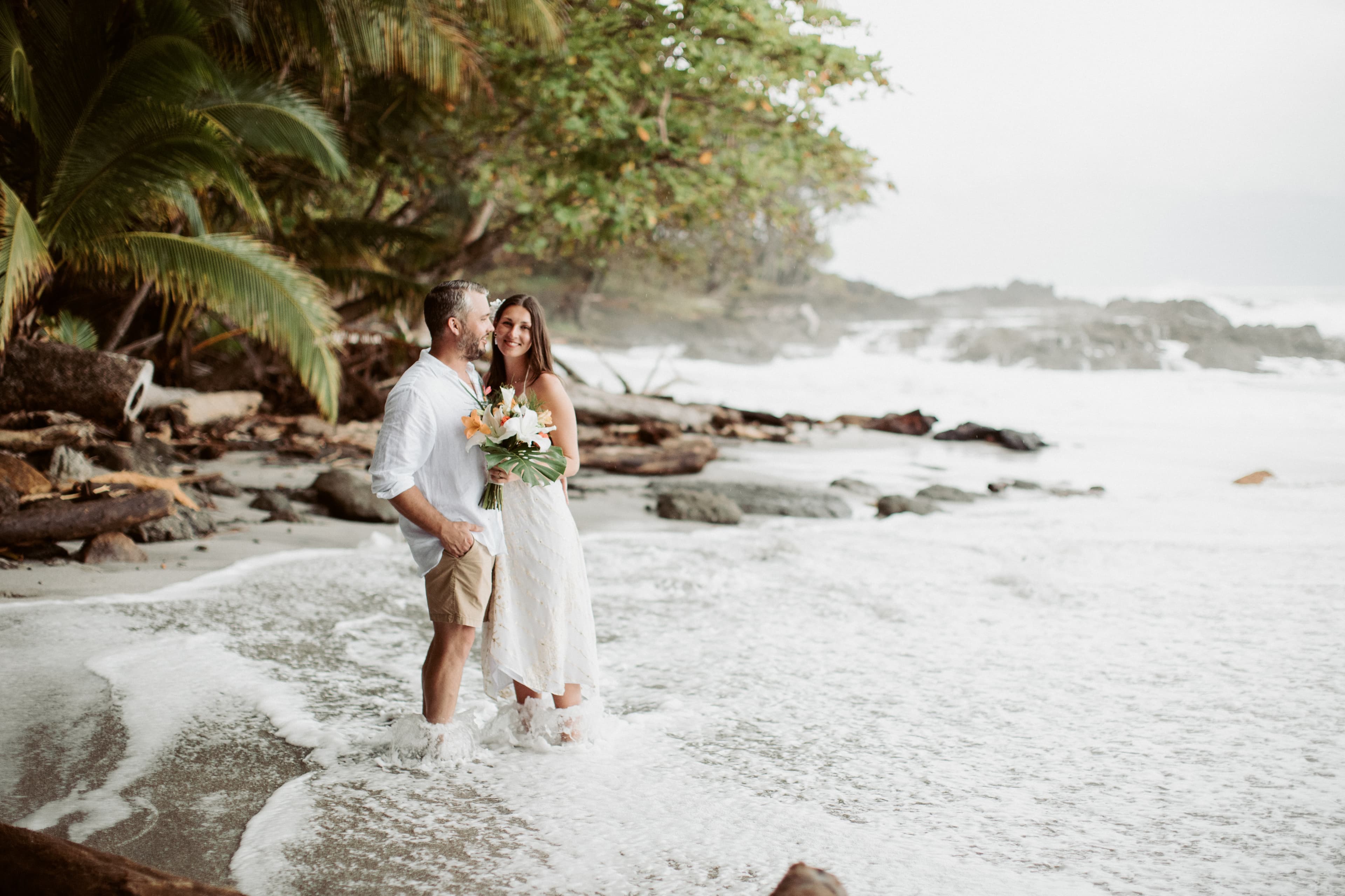 Bride and groom standing on a rocky tropical beach, the smiling bride is holding a bouquet of tropical flowers and the groom is wearing a loose white collared shirt and tan shorts and is facing the bride