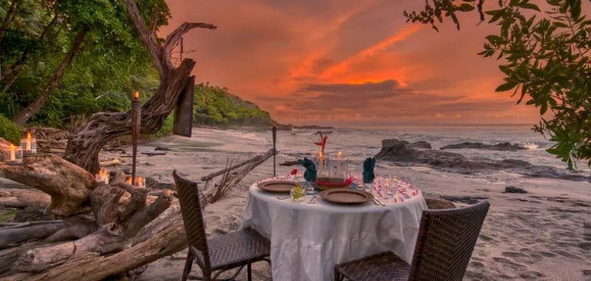 A romantic beachside dinner setup at sunset with a table adorned with candles and decorative tableware.