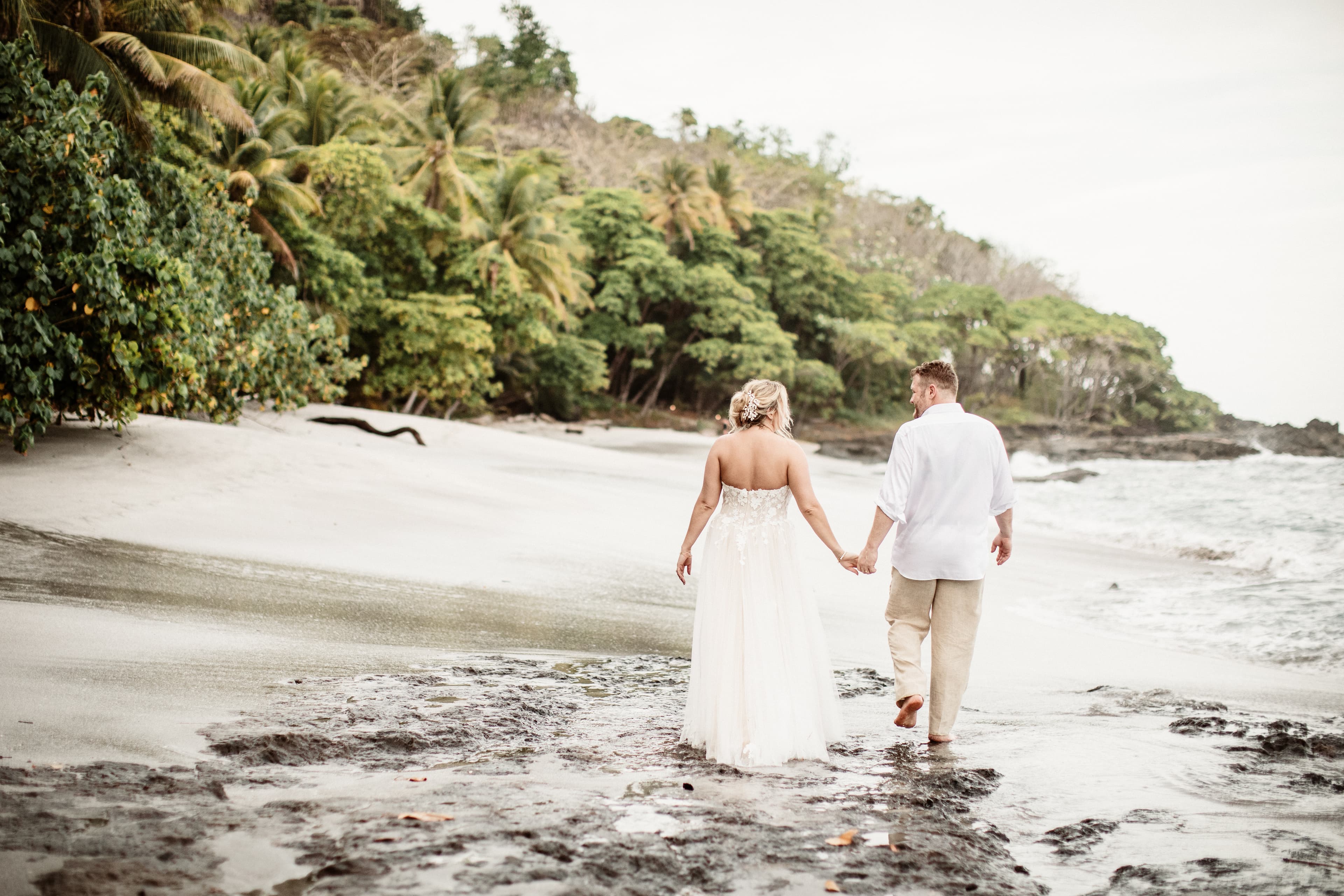 Bride and groom walking on the beach in wedding clothes holding hands.