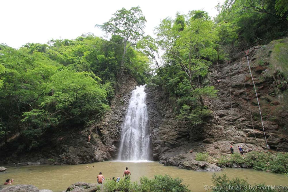 The view from the bottom of a waterfall looking up the rocky cliff with tropical vegetation