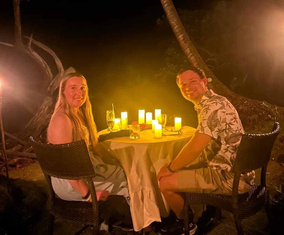 A couple sits at a candlelit table outdoors at night, smiling at the camera.