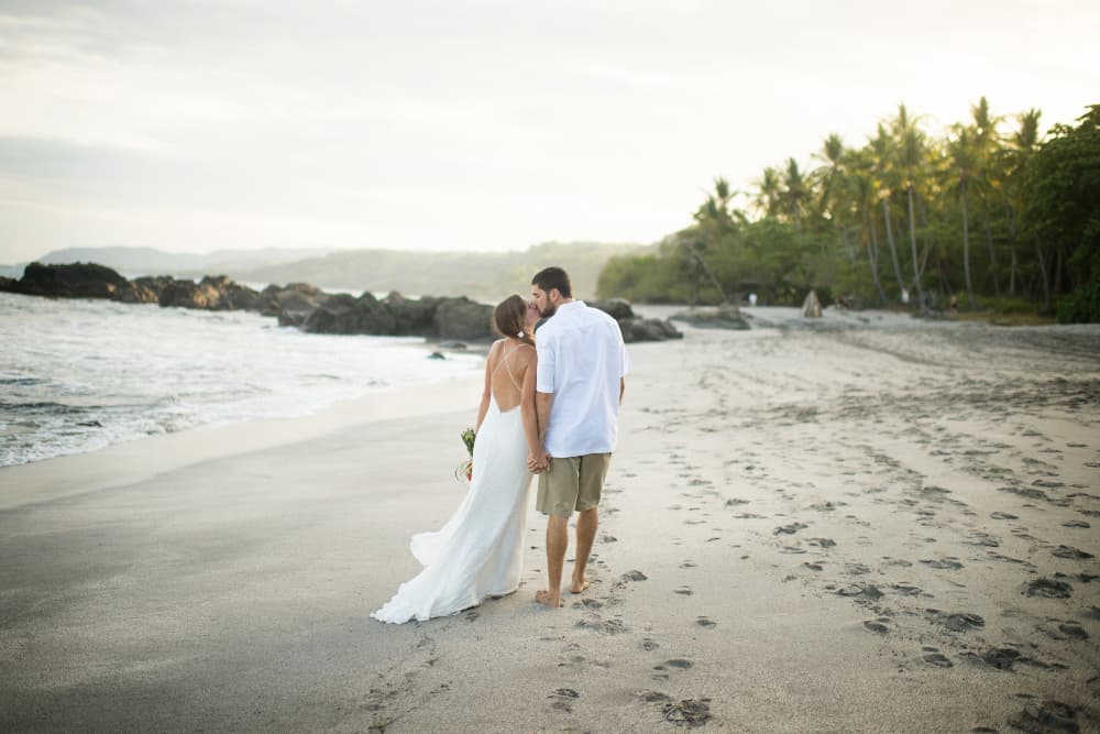 A wedding couple kissing on a sandy beach with the ocean and palm trees around them.
