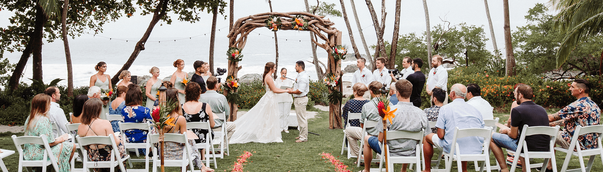 Bride and Group under driftwood arbor, ocean in the background, with attendees sitting on white chairs with flower petals lining center isle.